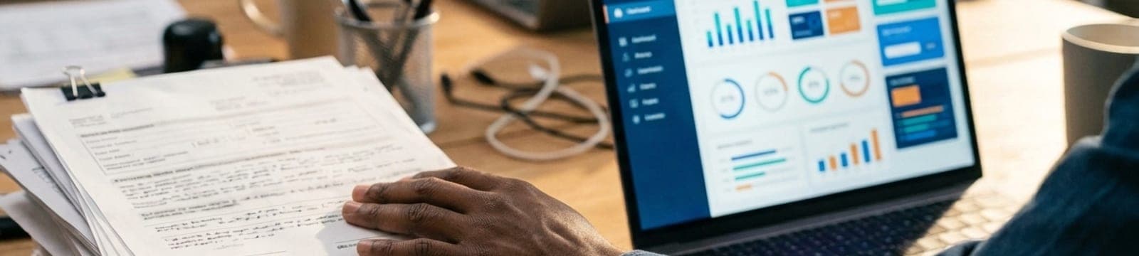 Hand pushing a stack of paper forms aside toward a laptop showing a dashboard